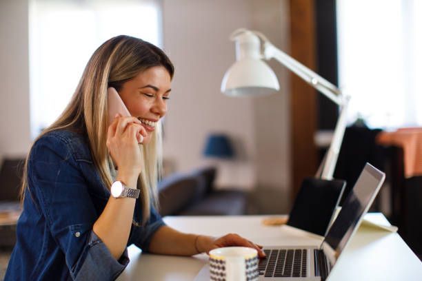 A woman is sitting at a desk talking on a cell phone while using a laptop computer.