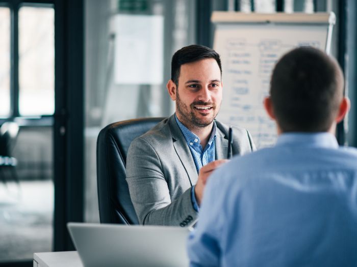 A man is sitting at a desk talking to another man.