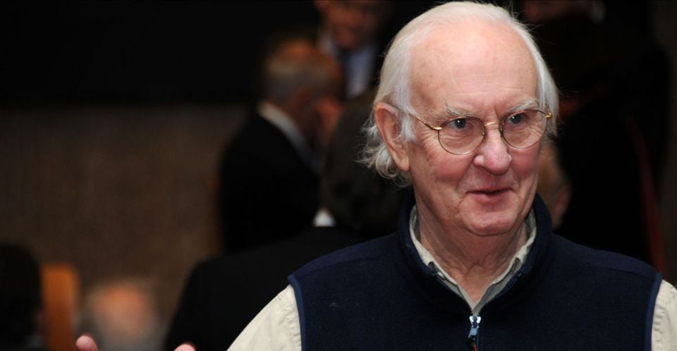 An older man wearing glasses and a vest is standing in a crowd.