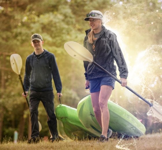 Two people with paddles stand near green kayaks outdoors with sunlight.