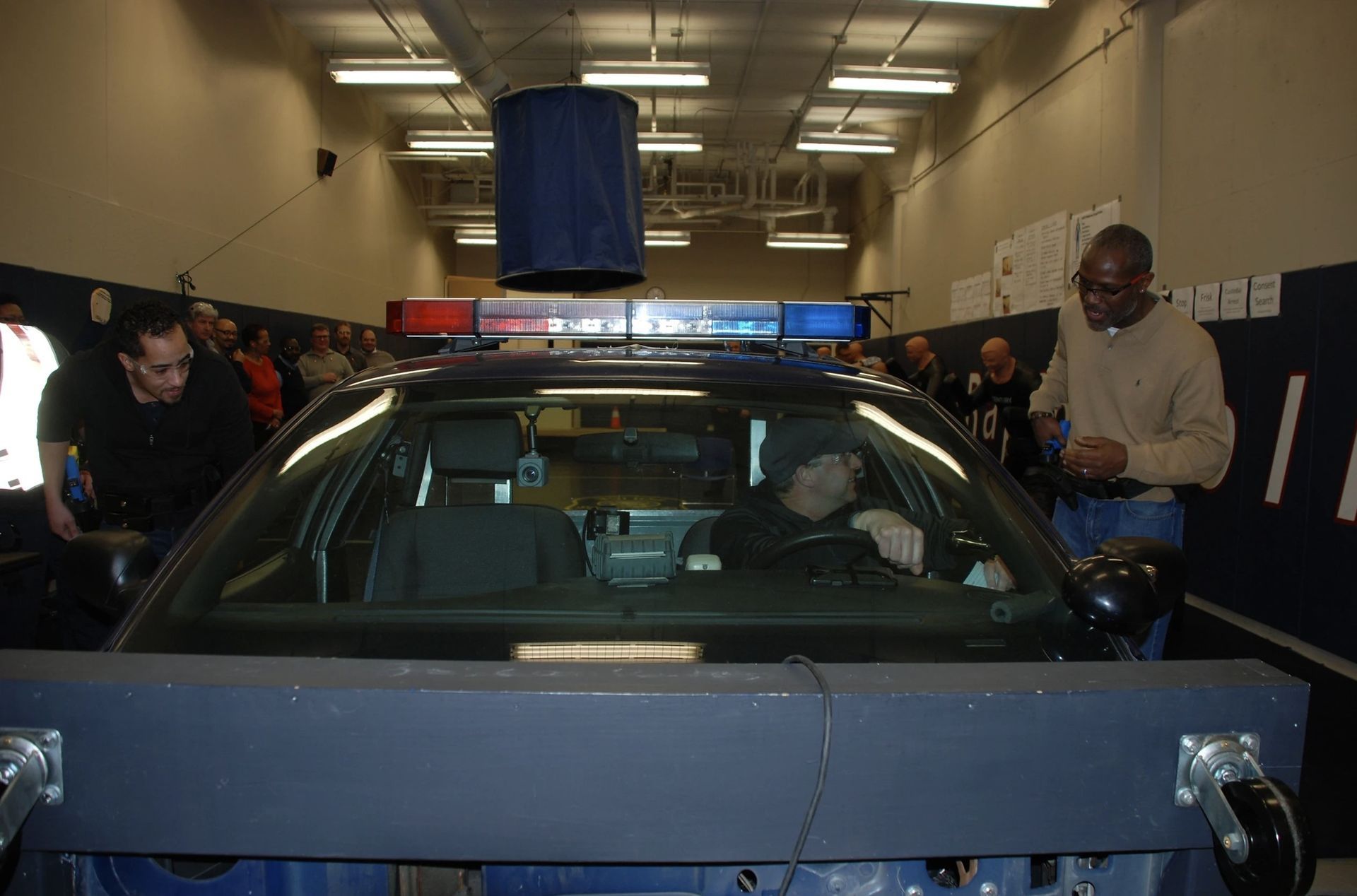 Police car interior, officers in driver's seat, surrounded by people, testing lights in a room.