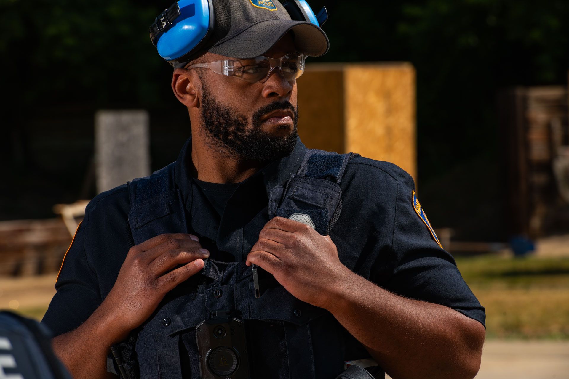 Man in tactical gear and ear protection at an outdoor shooting range.