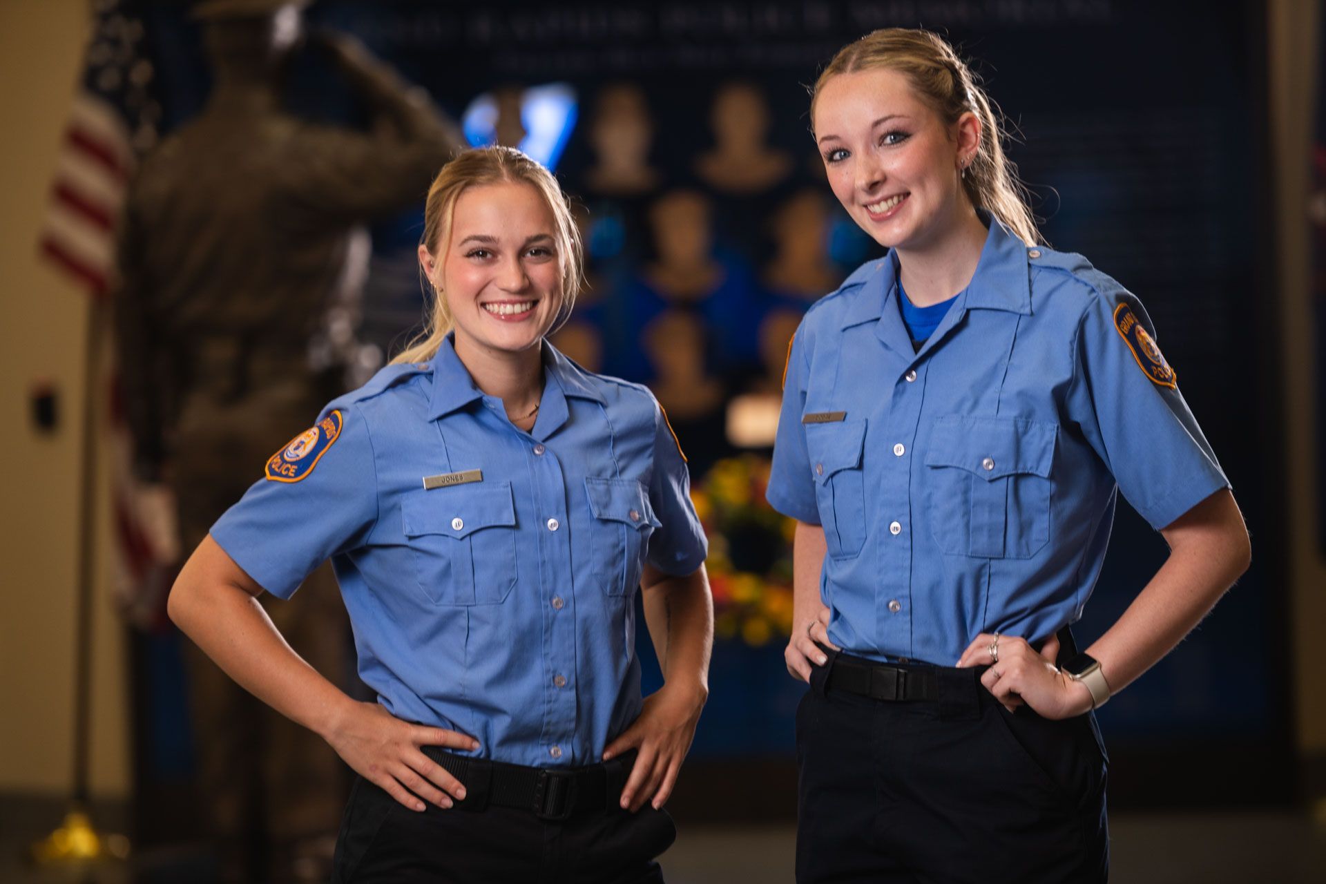Two women in blue uniform shirts and black pants smile, hands on hips, in front of a memorial.