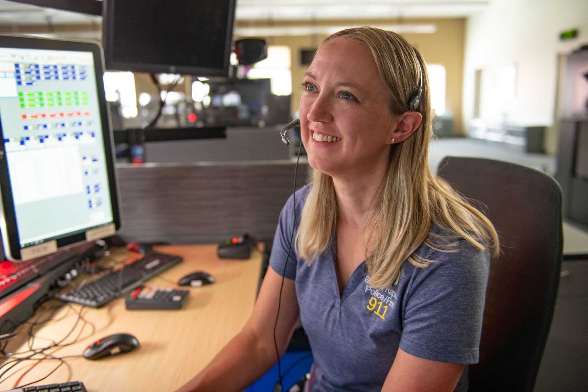 Woman with headset smiles at a computer, working in a 911 dispatch center.