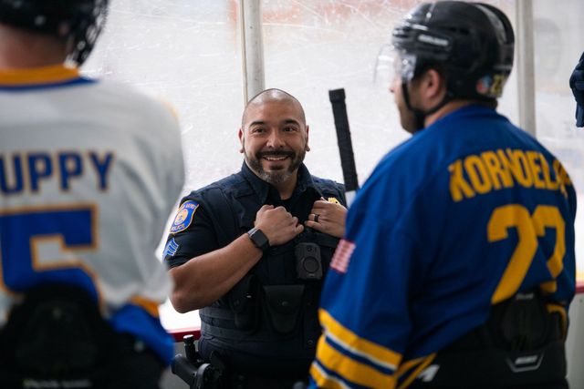 Smiling police officer in uniform poses between two hockey players on ice.