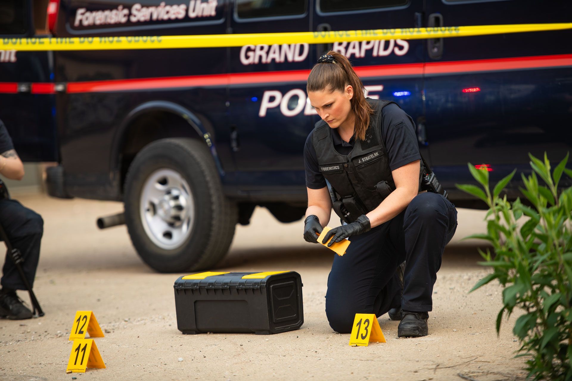 Police officer kneeling near evidence markers at a crime scene. A forensic van is in the background.