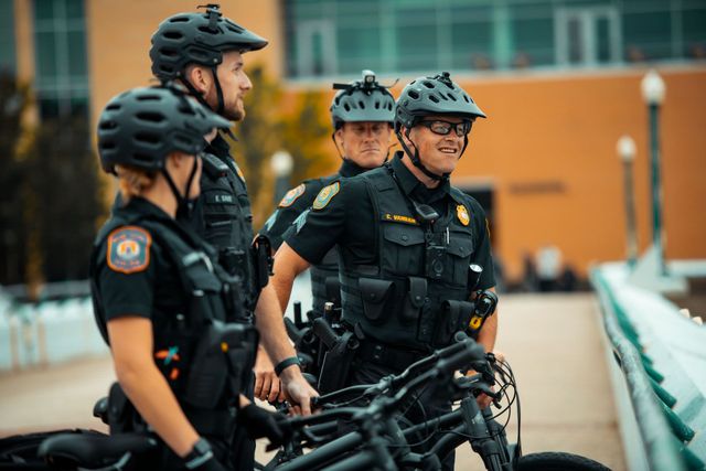 Four police officers in black uniforms and helmets stand near their bicycles on a bridge; one smiles.