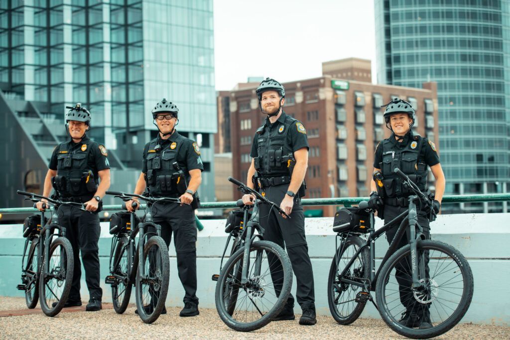 Four police officers on bicycles posing in front of urban buildings. They wear black uniforms and helmets.