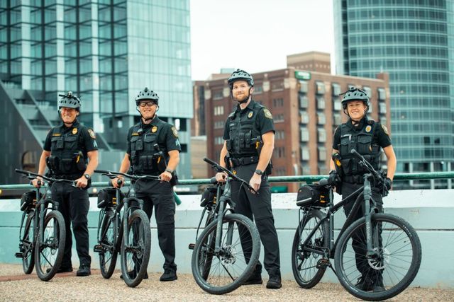 Four police officers on black bicycles pose in front of city buildings. They wear helmets, vests, and uniforms.