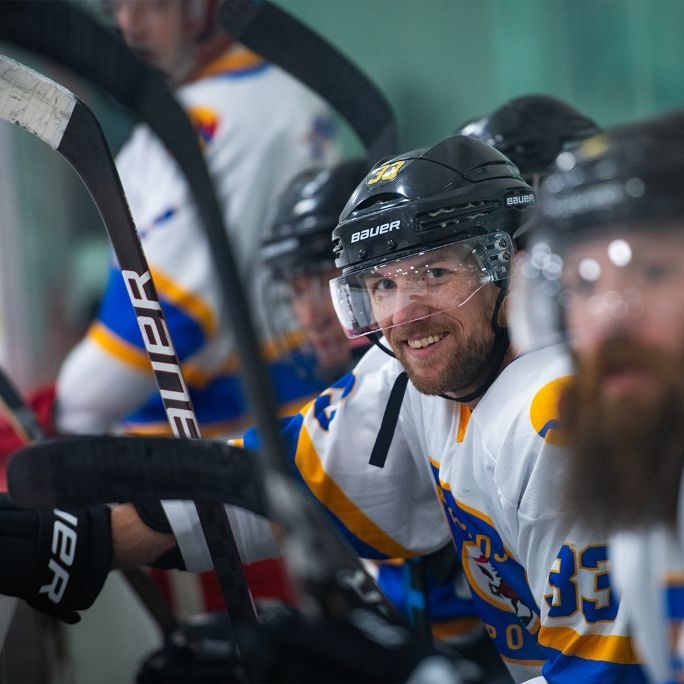 Hockey player in a white and blue jersey, smiling, wearing a helmet. Benches, other players, and hockey sticks are visible.