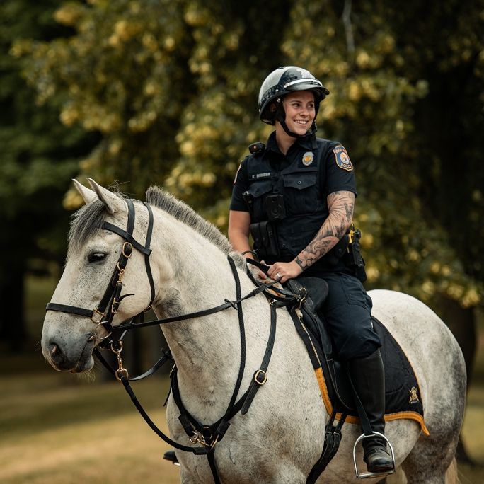 Police officer on horseback, wearing helmet, smiling, in front of a tree.