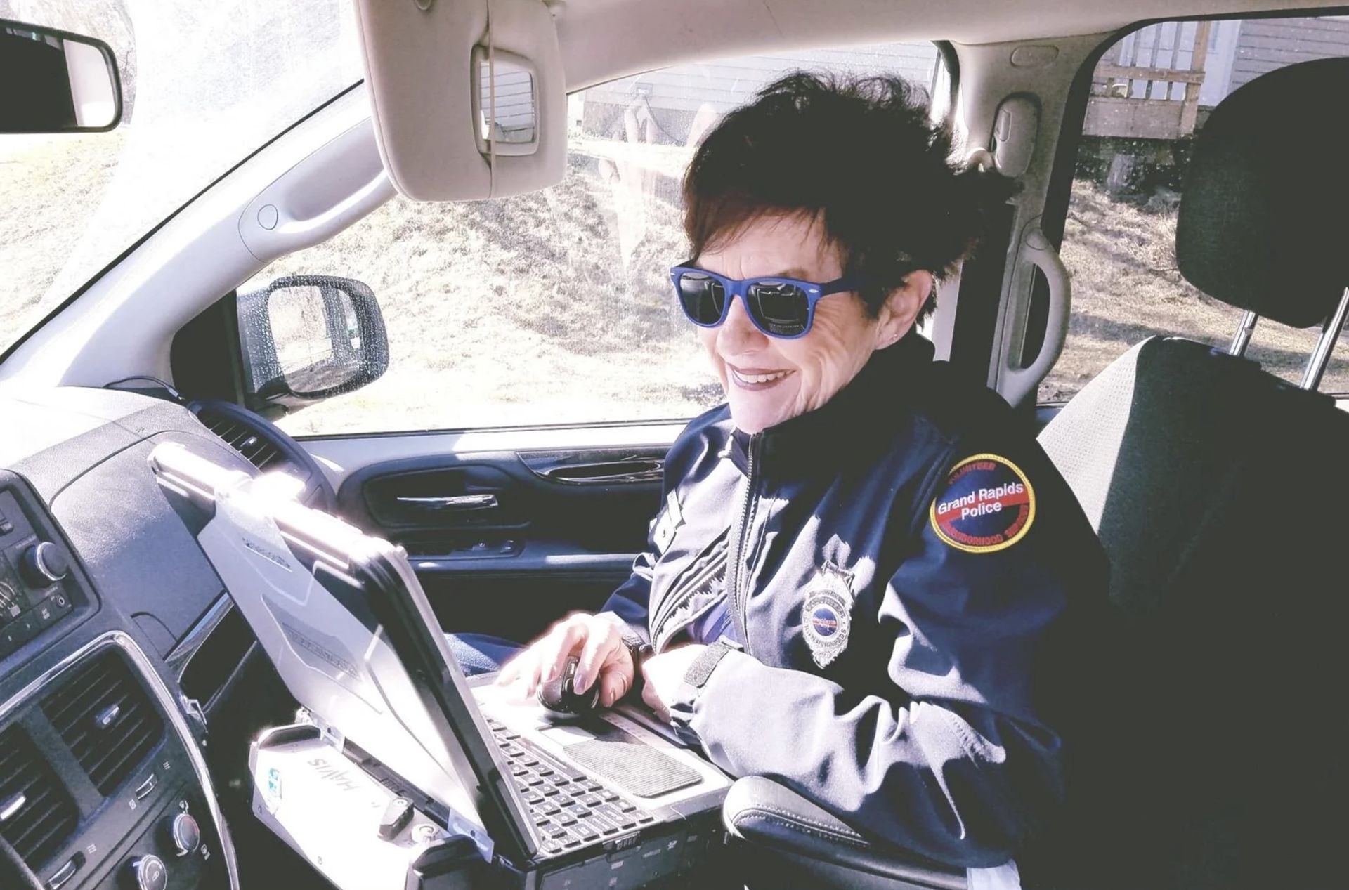 Woman in uniform, seated in a car, smiling while working on a laptop.