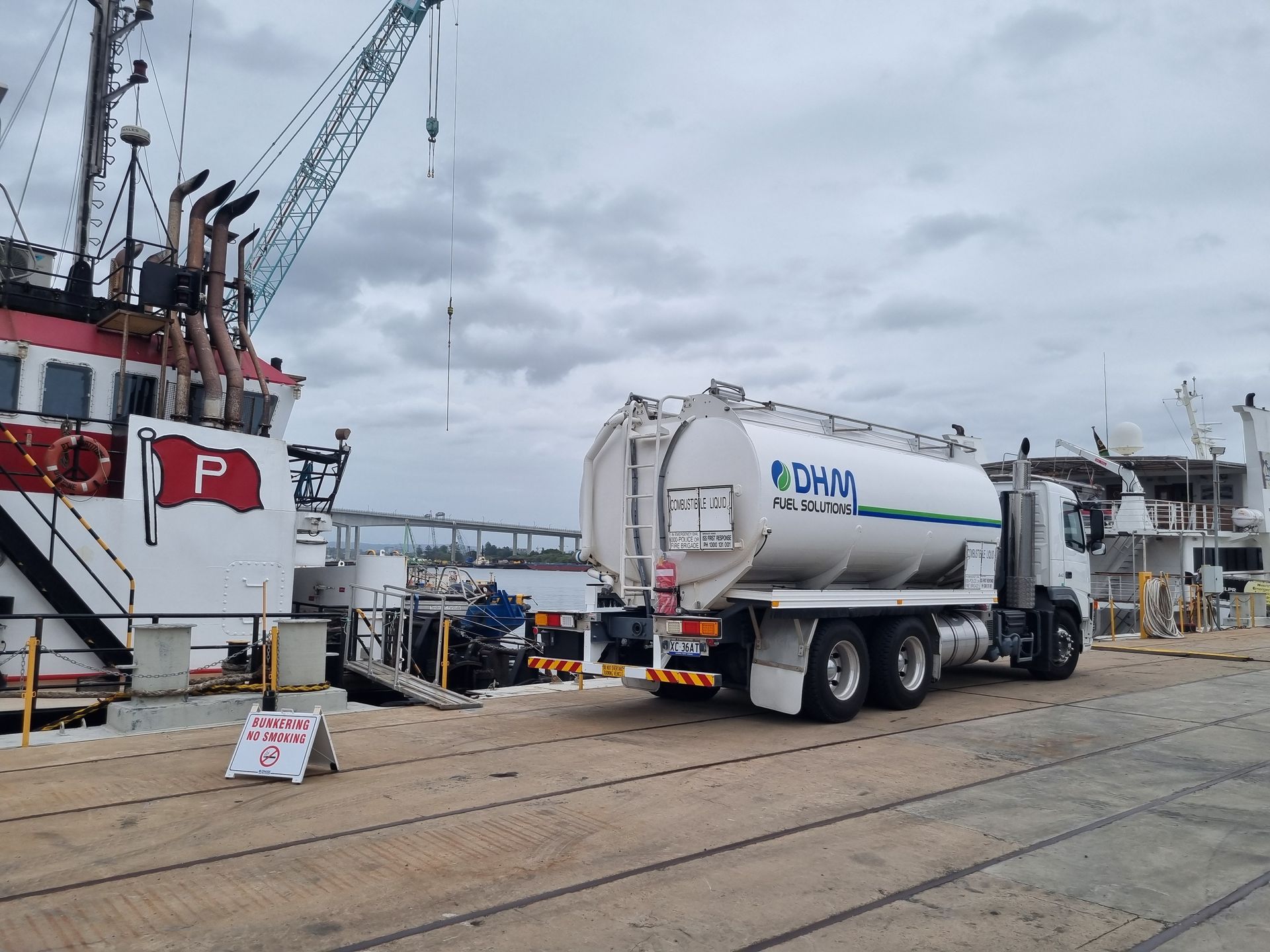 White tanker truck at a harbor dock beside a ship under gray skies.