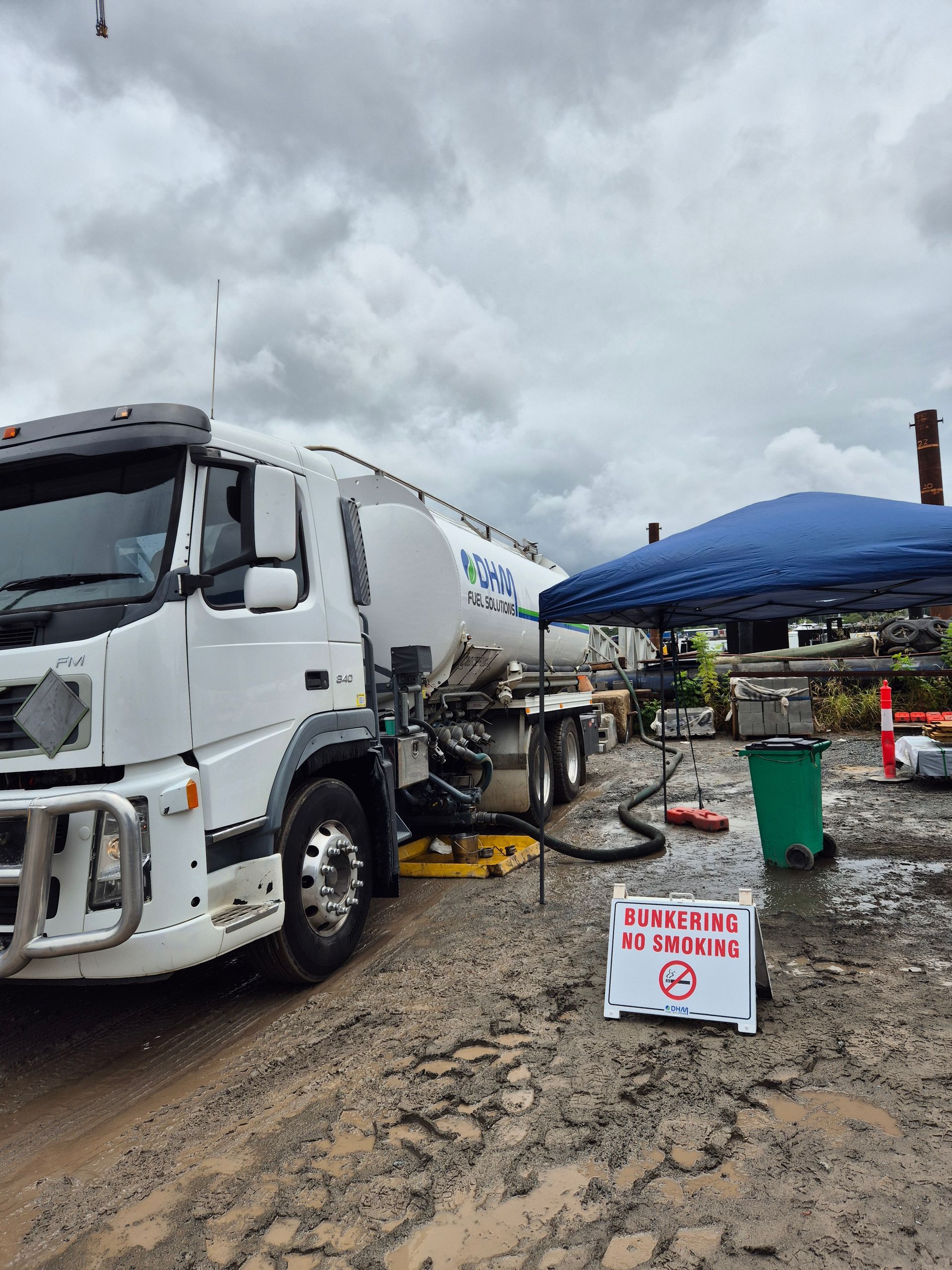 White tanker truck at a muddy roadside worksite beside a blue canopy and warning sign.