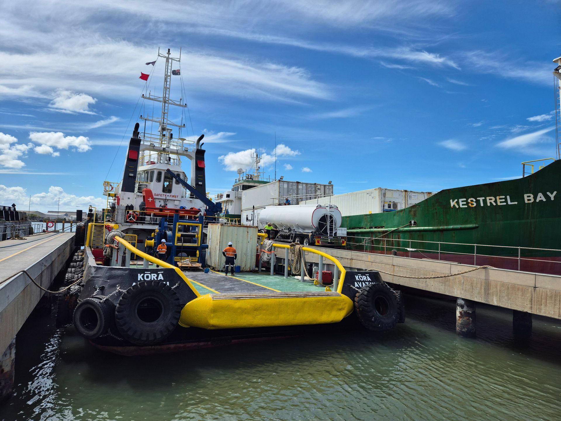 Yellow tugboat in a narrow harbor lock beside a large green cargo ship under a blue sky
