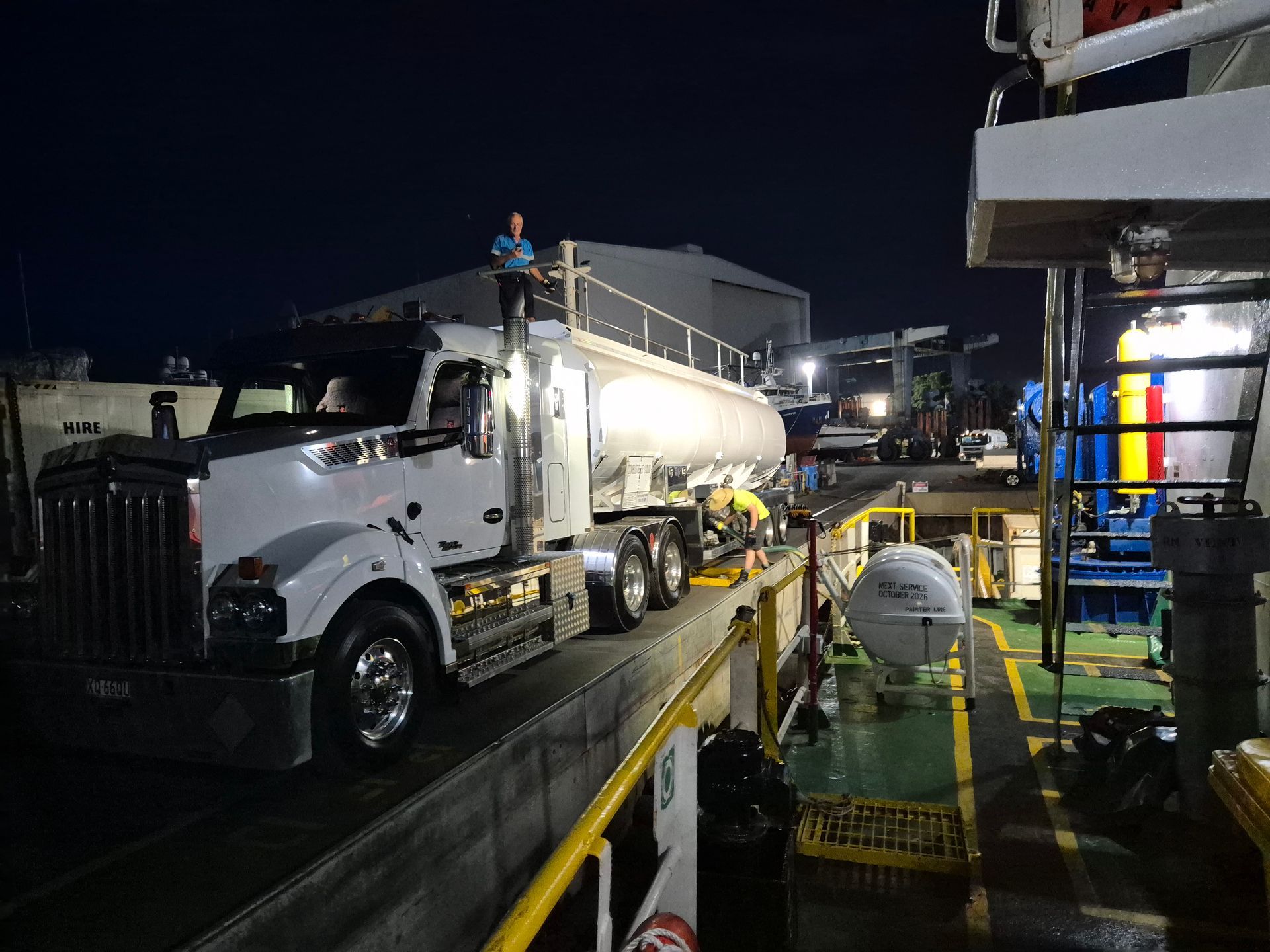 White tanker truck at a nighttime industrial site, with a worker standing on top near loading equipment.