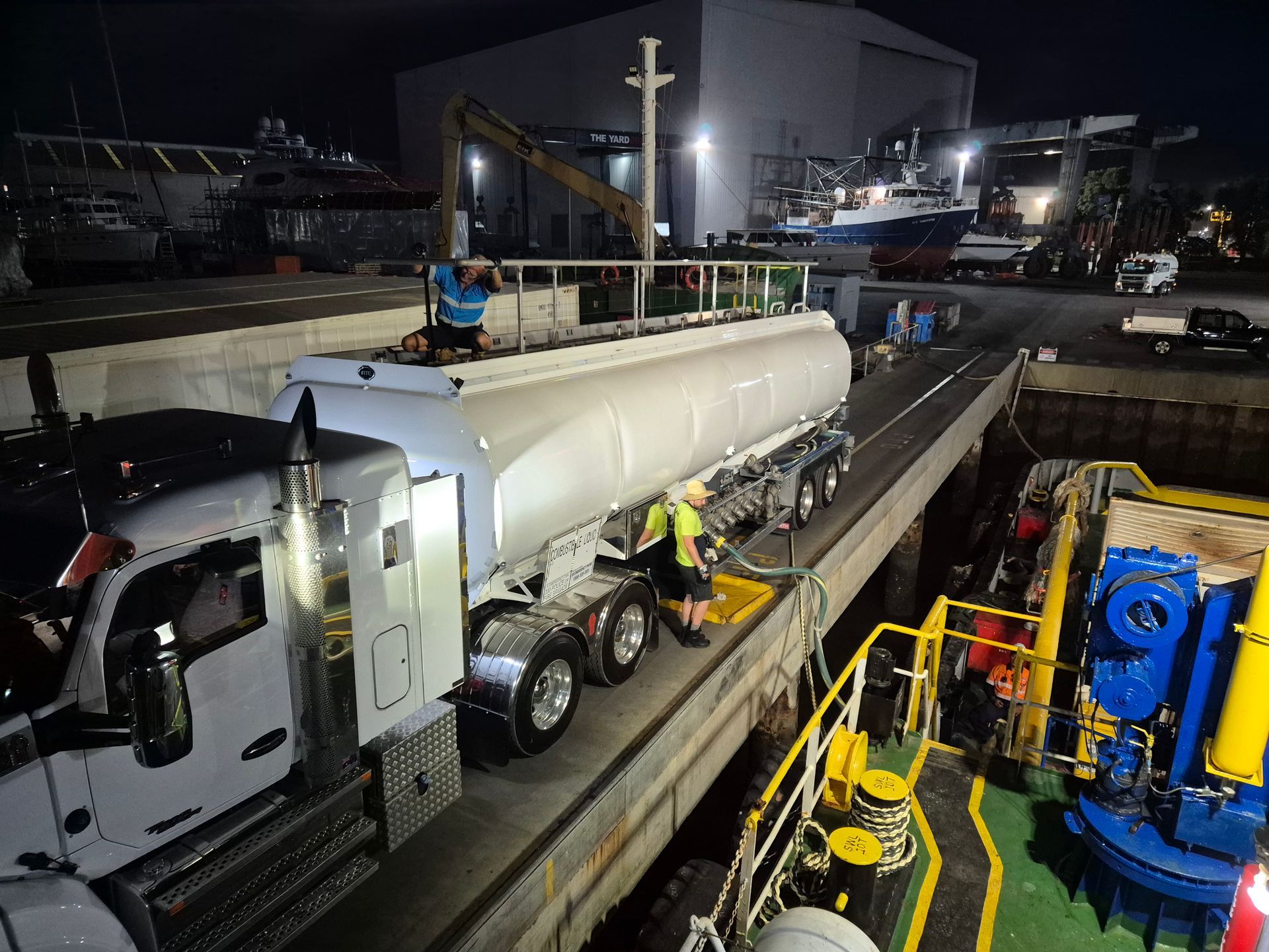 White tanker truck at night in an industrial yard, with workers inspecting it near heavy machinery