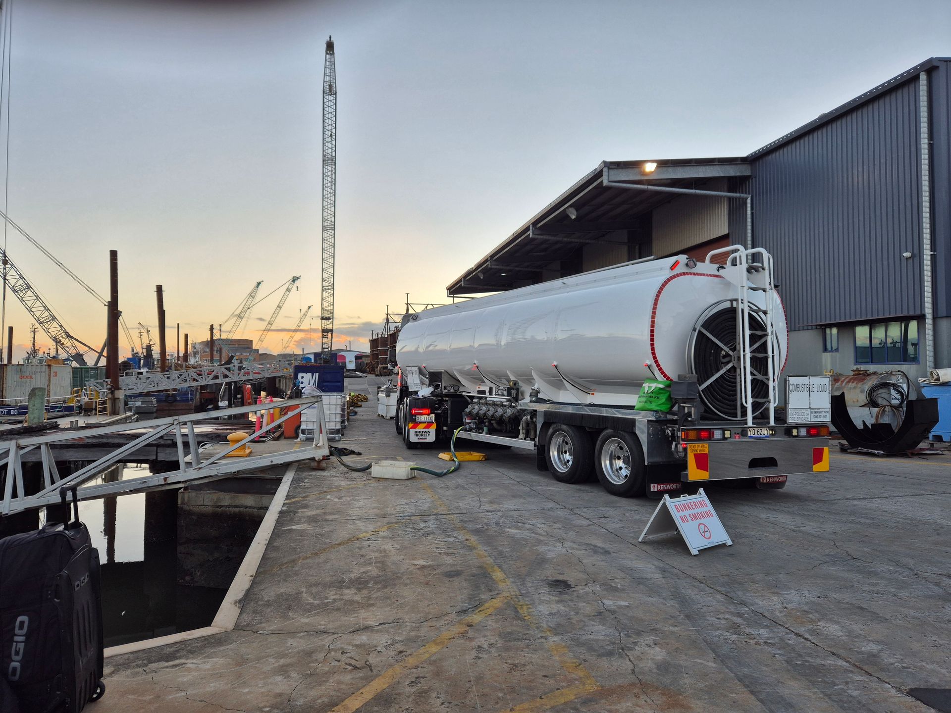 White tanker truck parked at a dockyard at sunset, with cranes and industrial buildings in the background