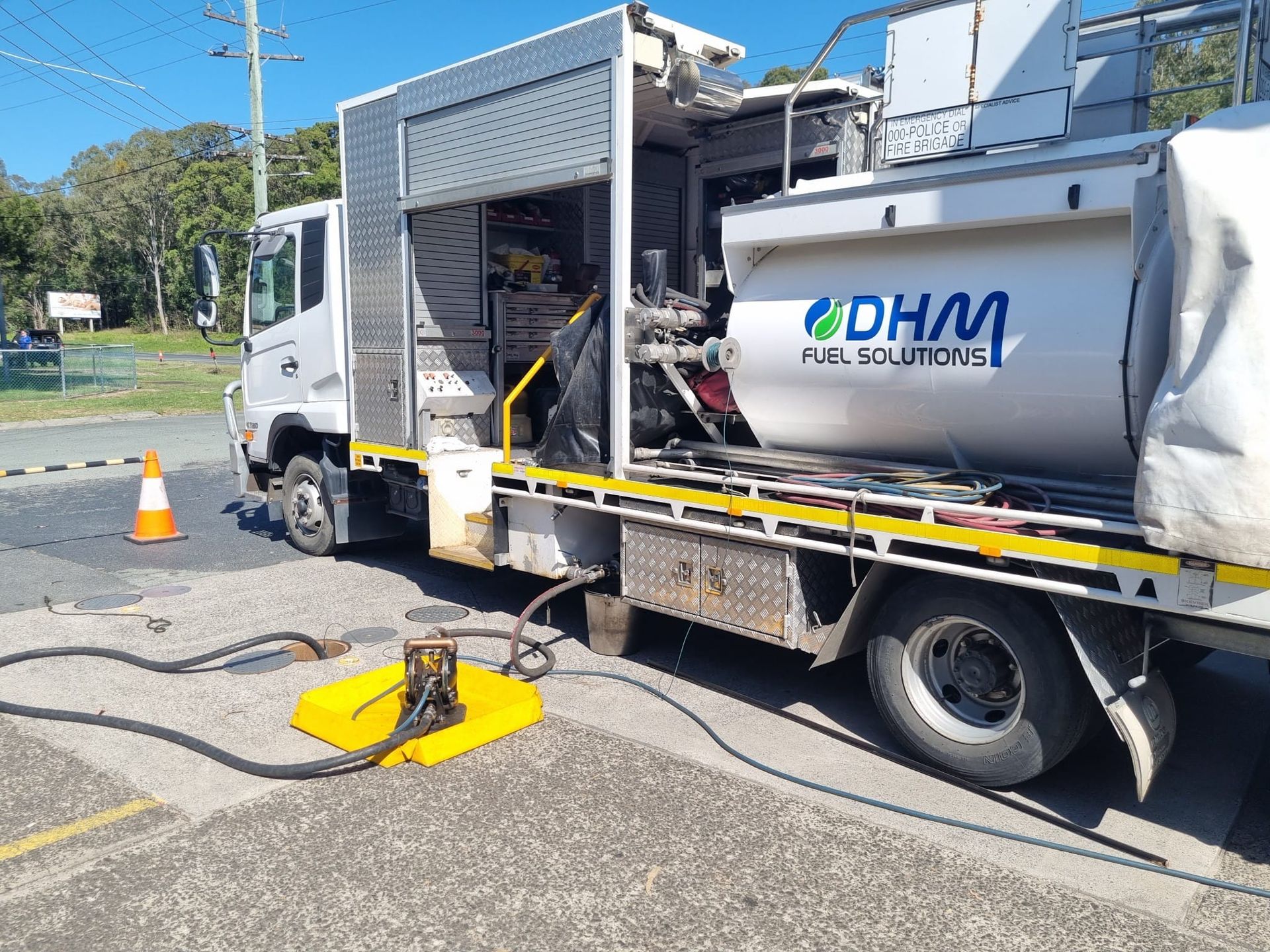 Fuel truck with open bay, refuelling at a roadside site. White truck with a large tank and hoses.