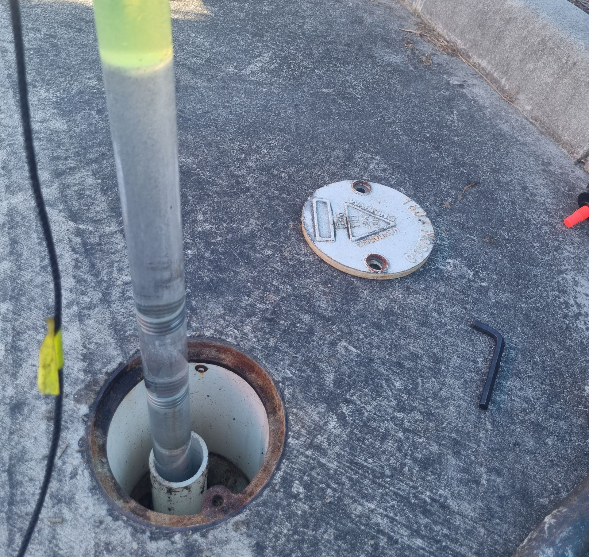 Yellow-sleeved pole in a pavement hole beside a curb with a round cover on asphalt.