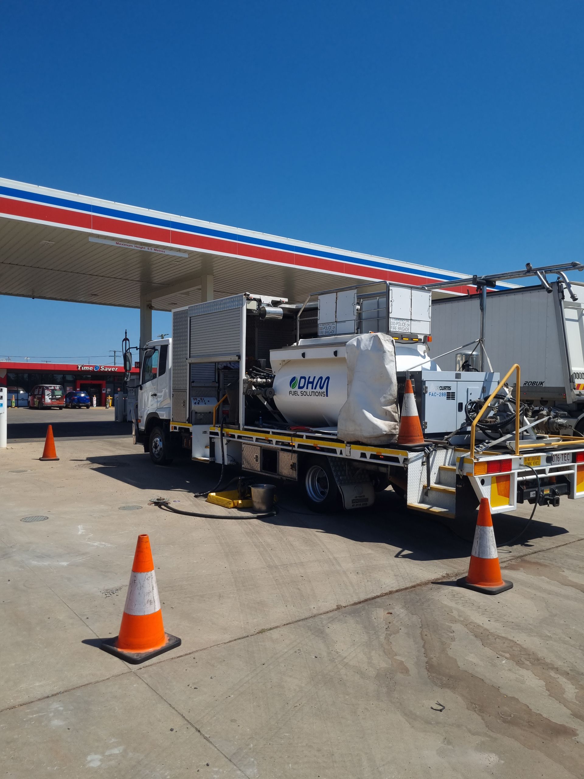 Truck parked at a gas station, equipped with machinery, orange cones present. Bright blue sky.