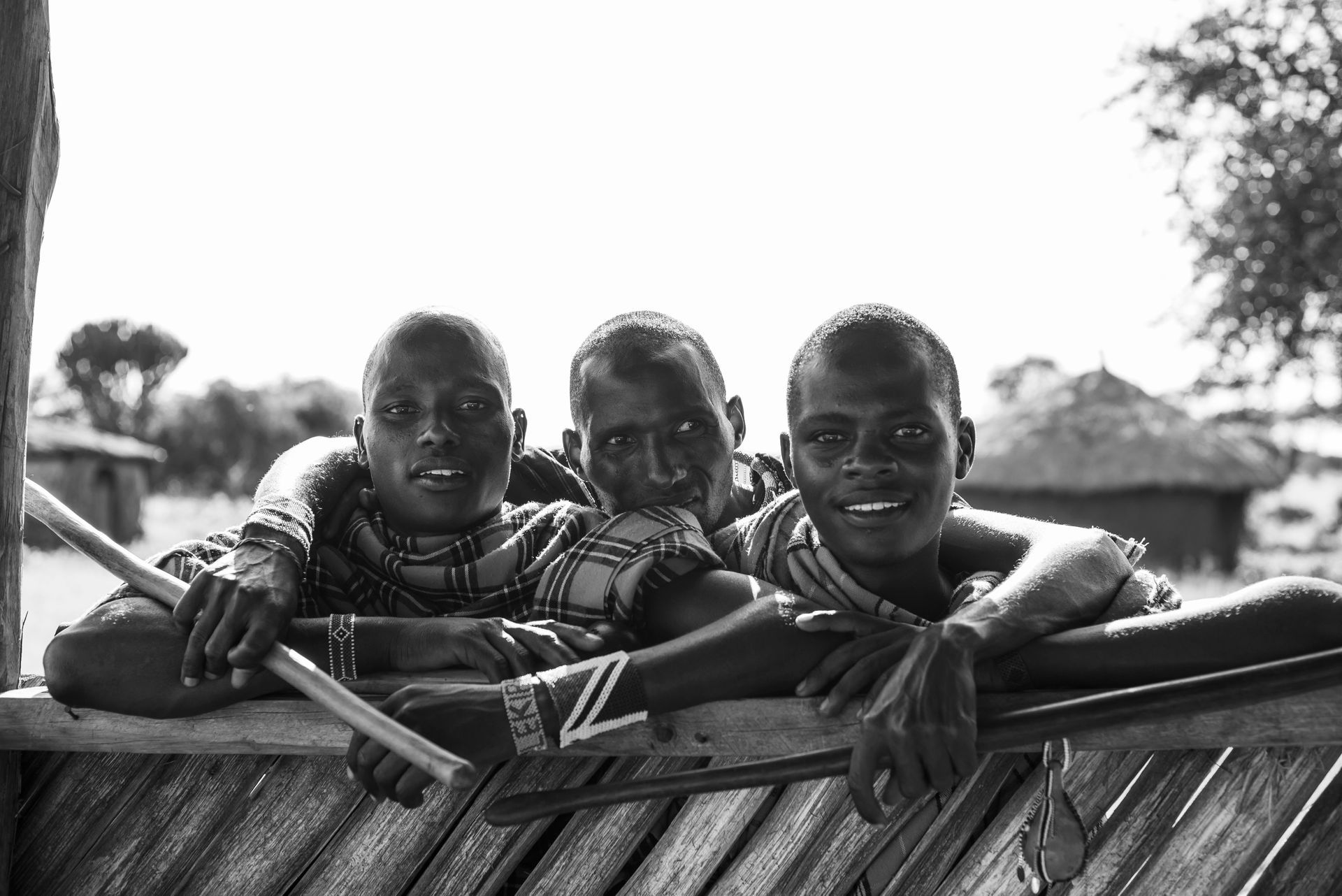 Three children leaning on a woven fence, smiling at the camera outdoors.