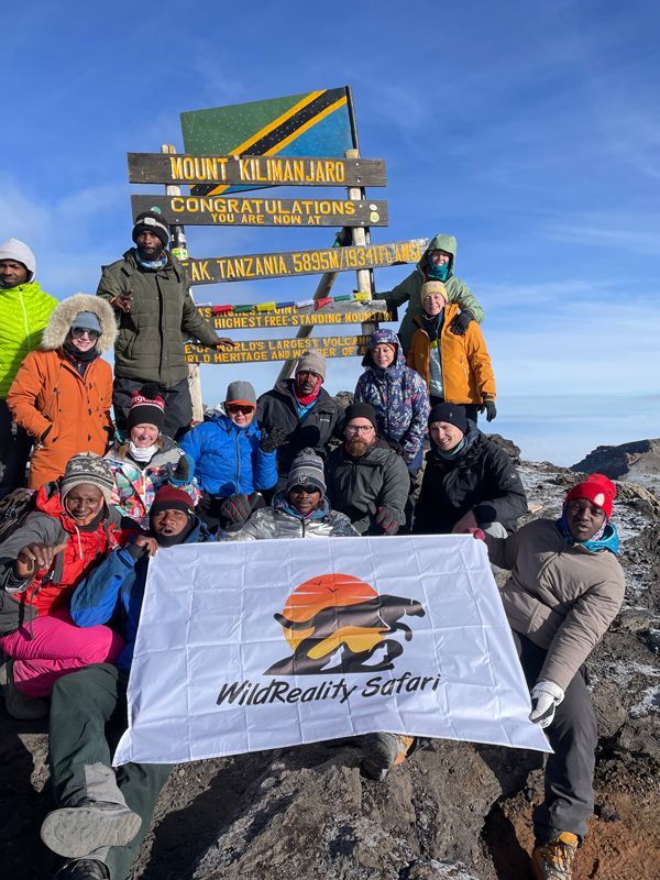 Group posing at Mount Kilimanjaro summit sign with a WildReality Safari banner in snowy conditions
