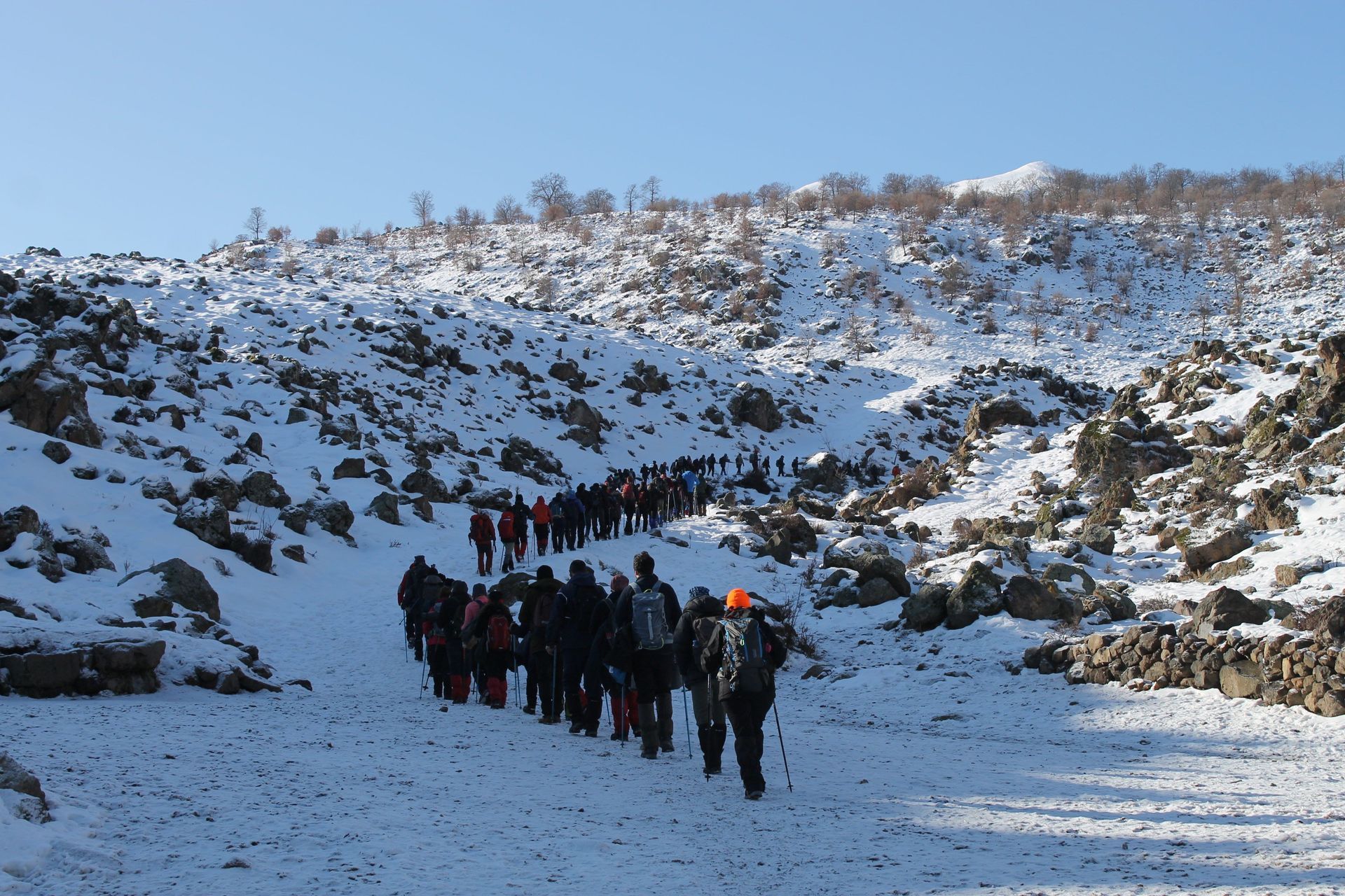 Hikers walking single file through a snowy mountain pass with rocky slopes and blue sky