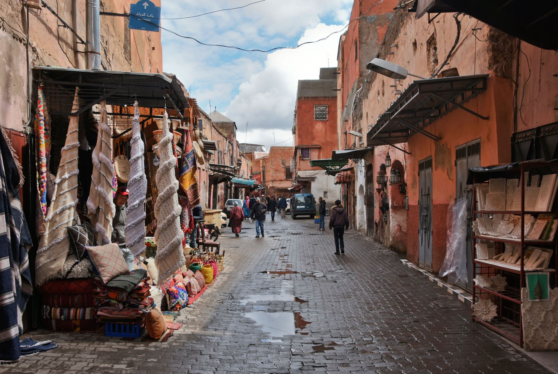 Narrow wet market street with colorful stalls, hanging goods, and a few people walking under cloudy sky
