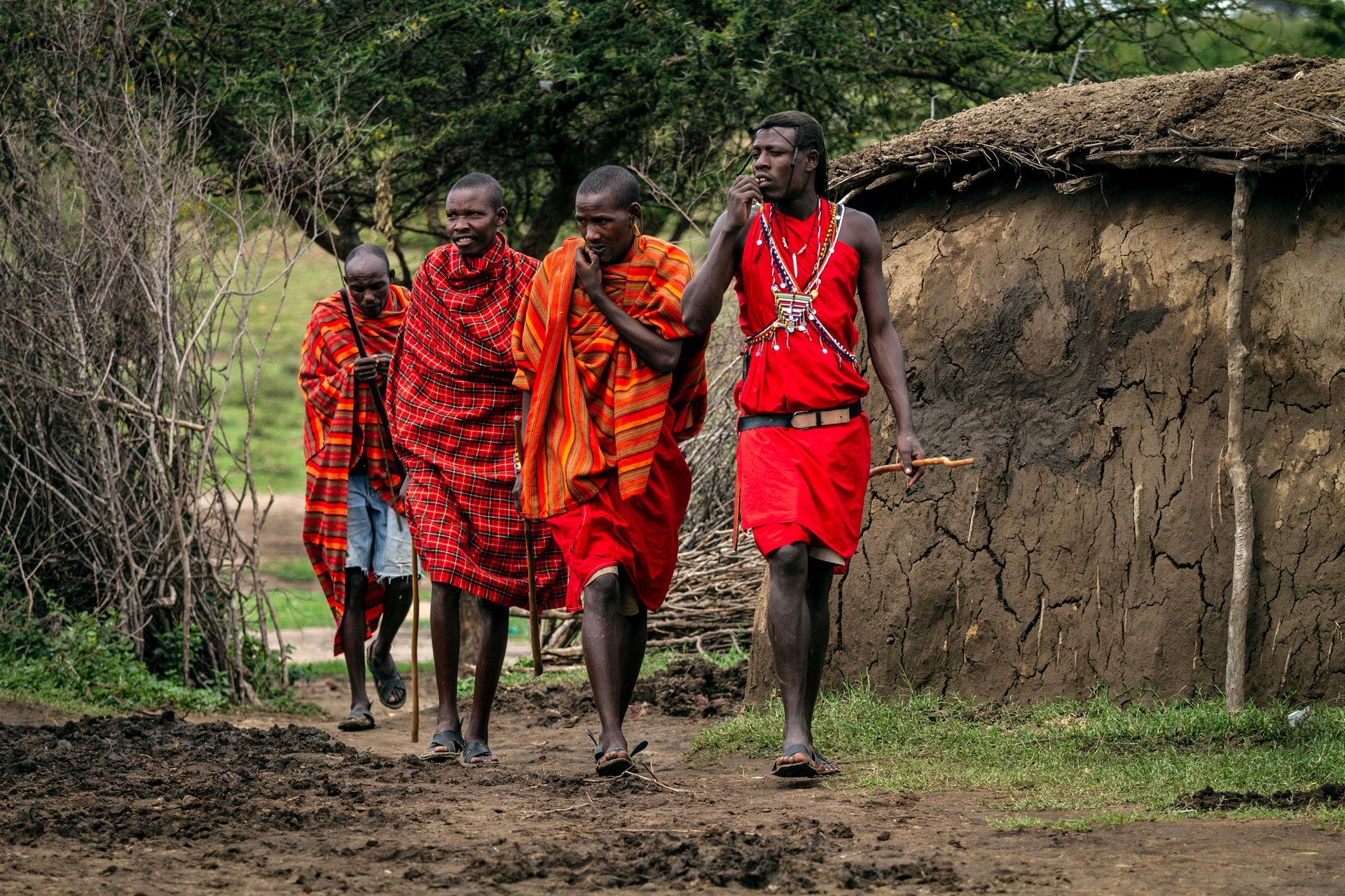 Four people in bright red and orange clothing walk along a muddy path beside a hut in a rural village.