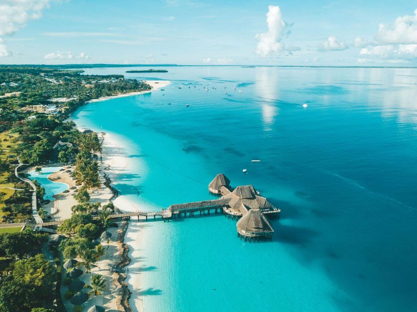 Aerial view of a tropical beach with turquoise water, palm trees, and overwater huts along the shore