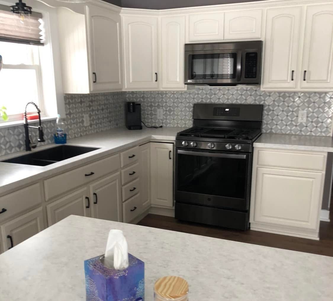 Modern kitchen with white cabinets, black stove and microwave, gray backsplash, and a window over the sink.