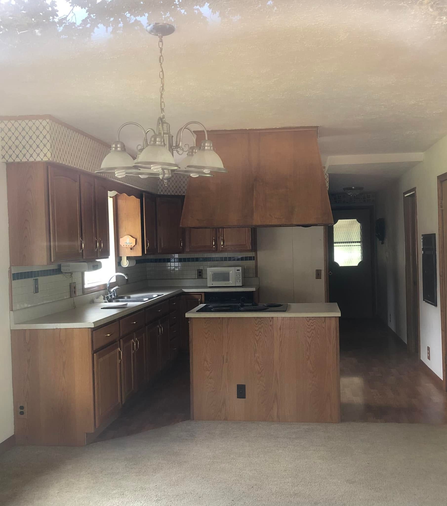 Small kitchen and dining area with brown cabinets, hanging light, and a hallway leading to a door.