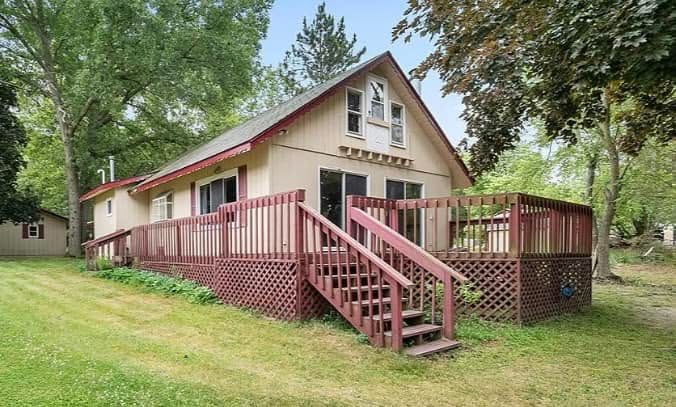 Small beige house with red trim, deck, and stairs on a grassy, tree-lined lot