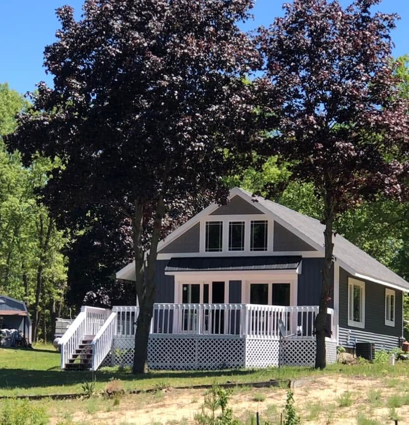 Gray house with white trim and porch under tall trees on a sunny lawn