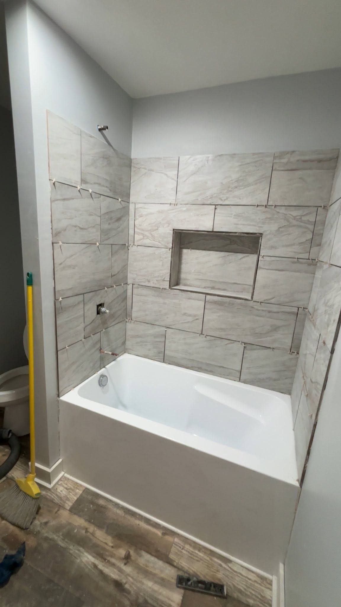 Bathroom alcove with a white tub, gray tile surround, and recessed shelf niche