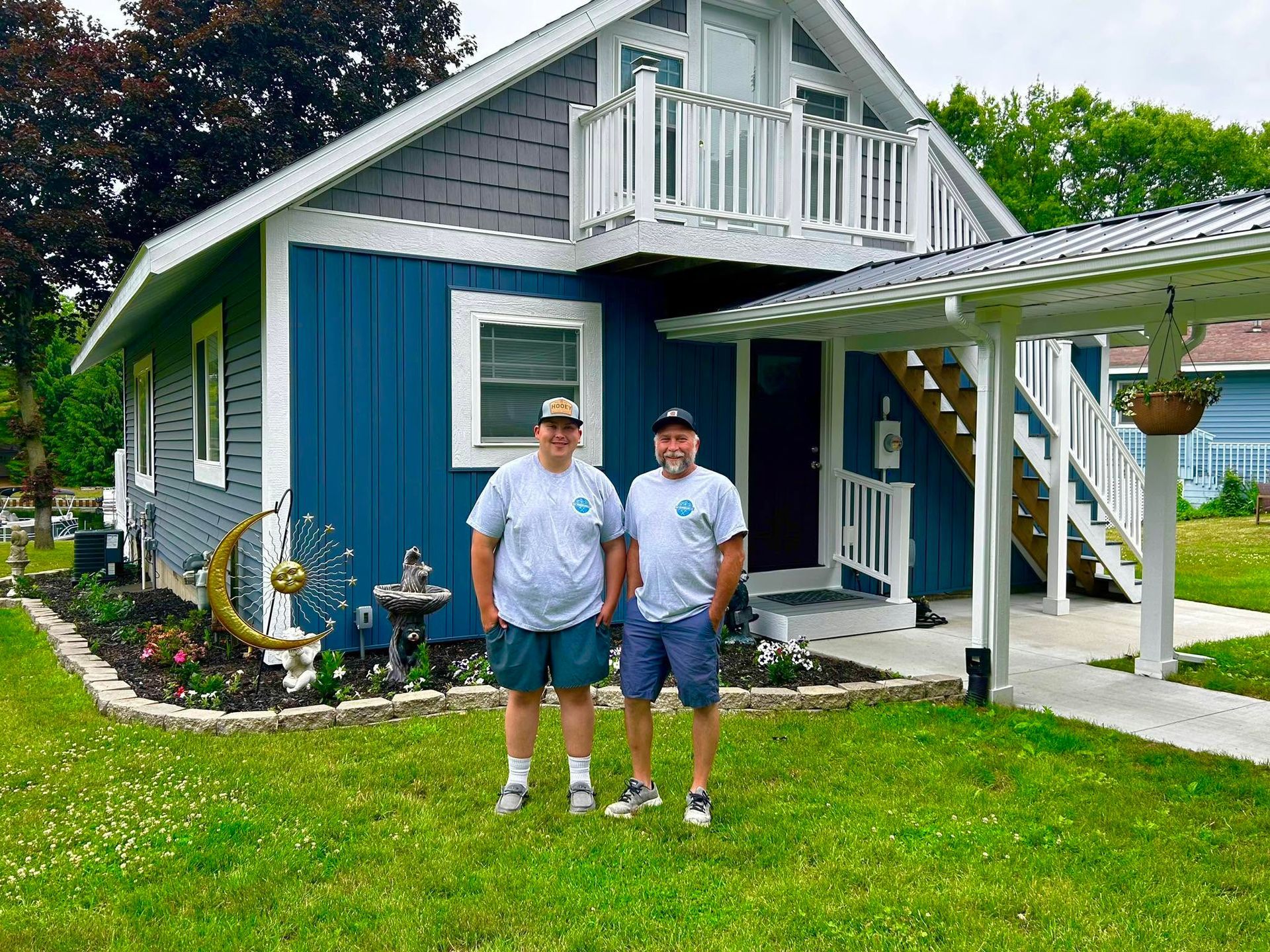 Two people standing on a lawn in front of a gray house with blue trim and a white porch.