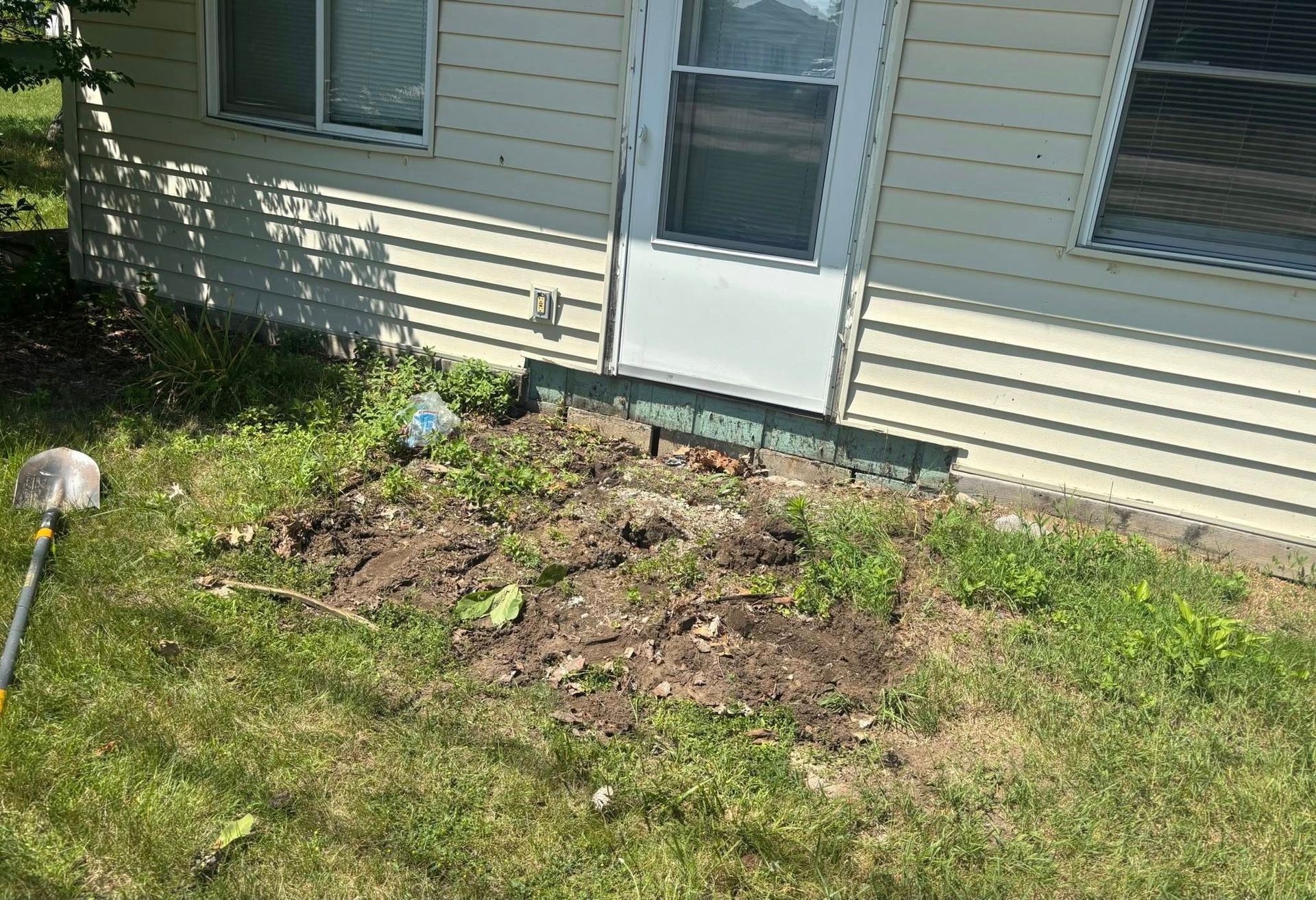 Patch of disturbed dirt and mulch beneath a house window, bordered by grass and a white door.