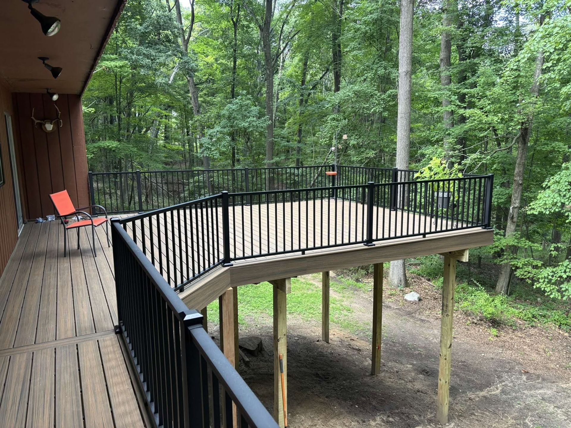 Wooden deck with black railing beside a forested house, extending over a sloped yard