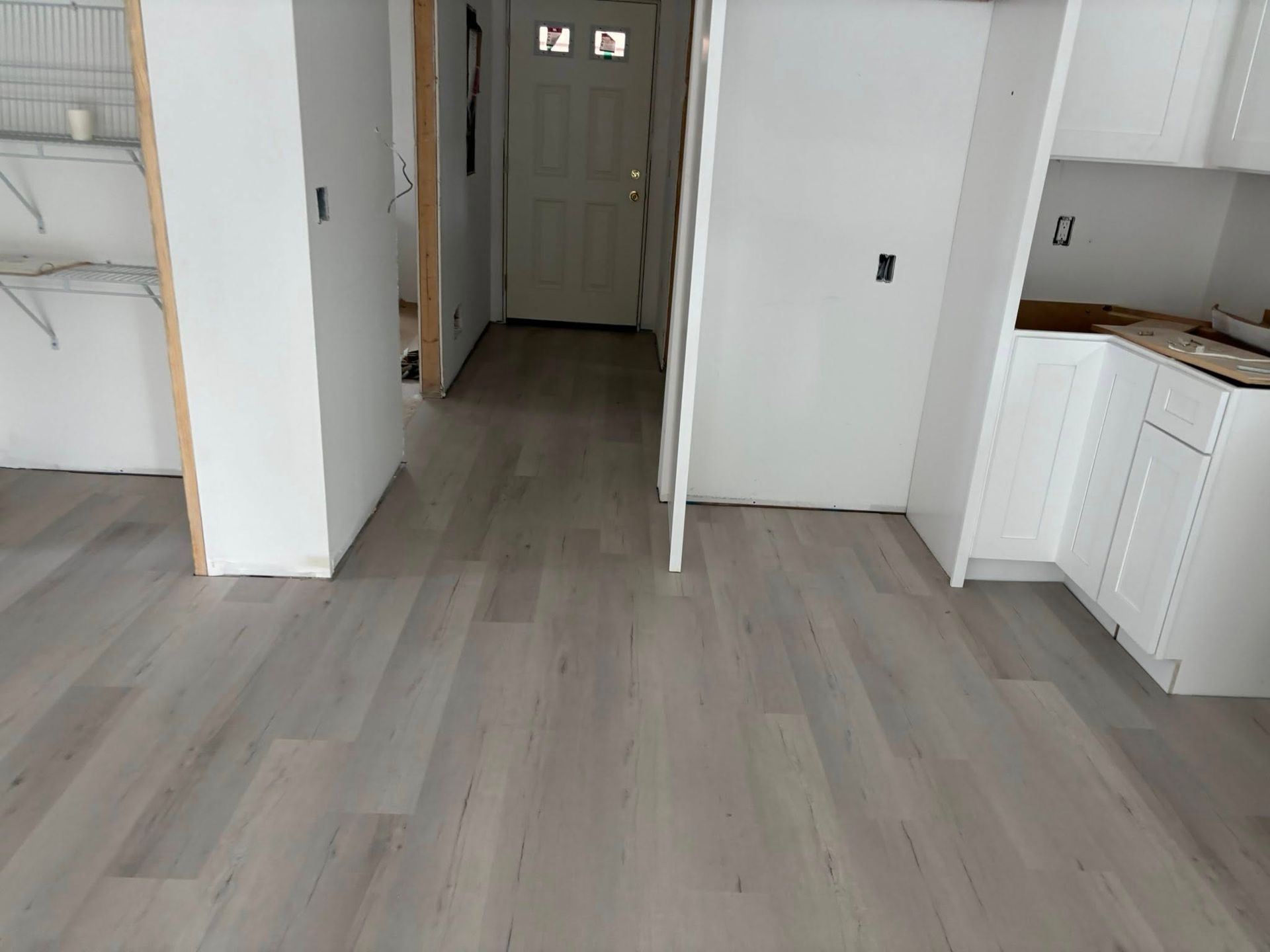 Empty modern room with light wood floors, white walls, and a hallway leading to a front door