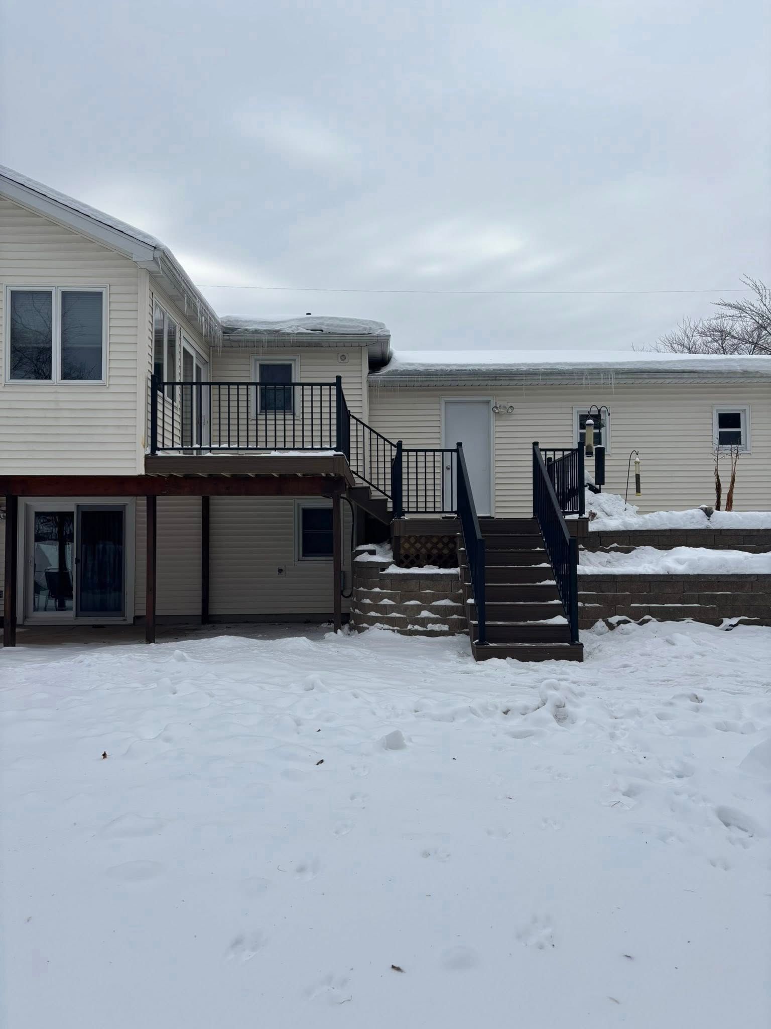 Snow-covered backyard with a two-story house and exterior stairs leading to a raised deck.