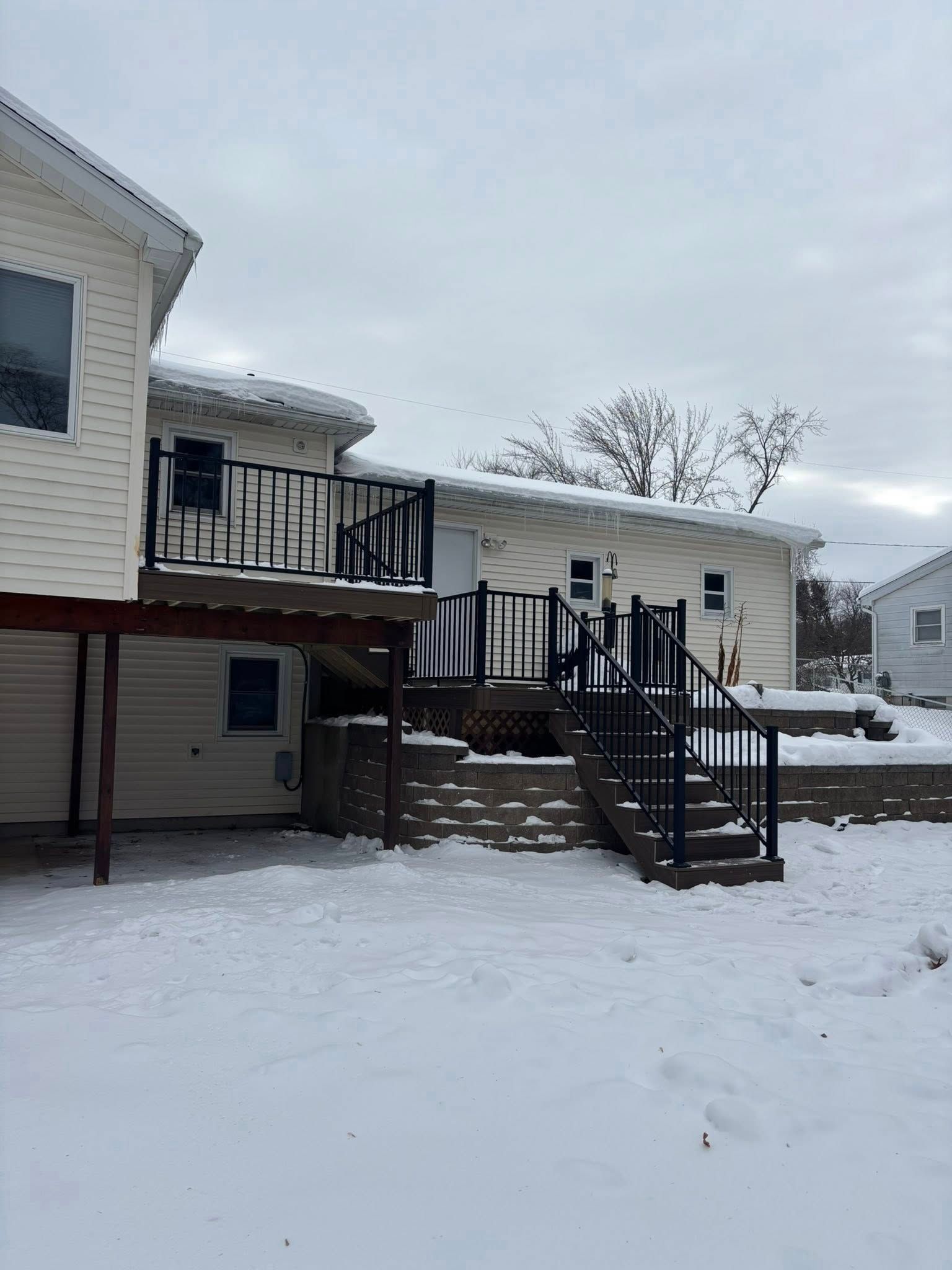Snowy backyard deck with stairs and black railings beside a beige house