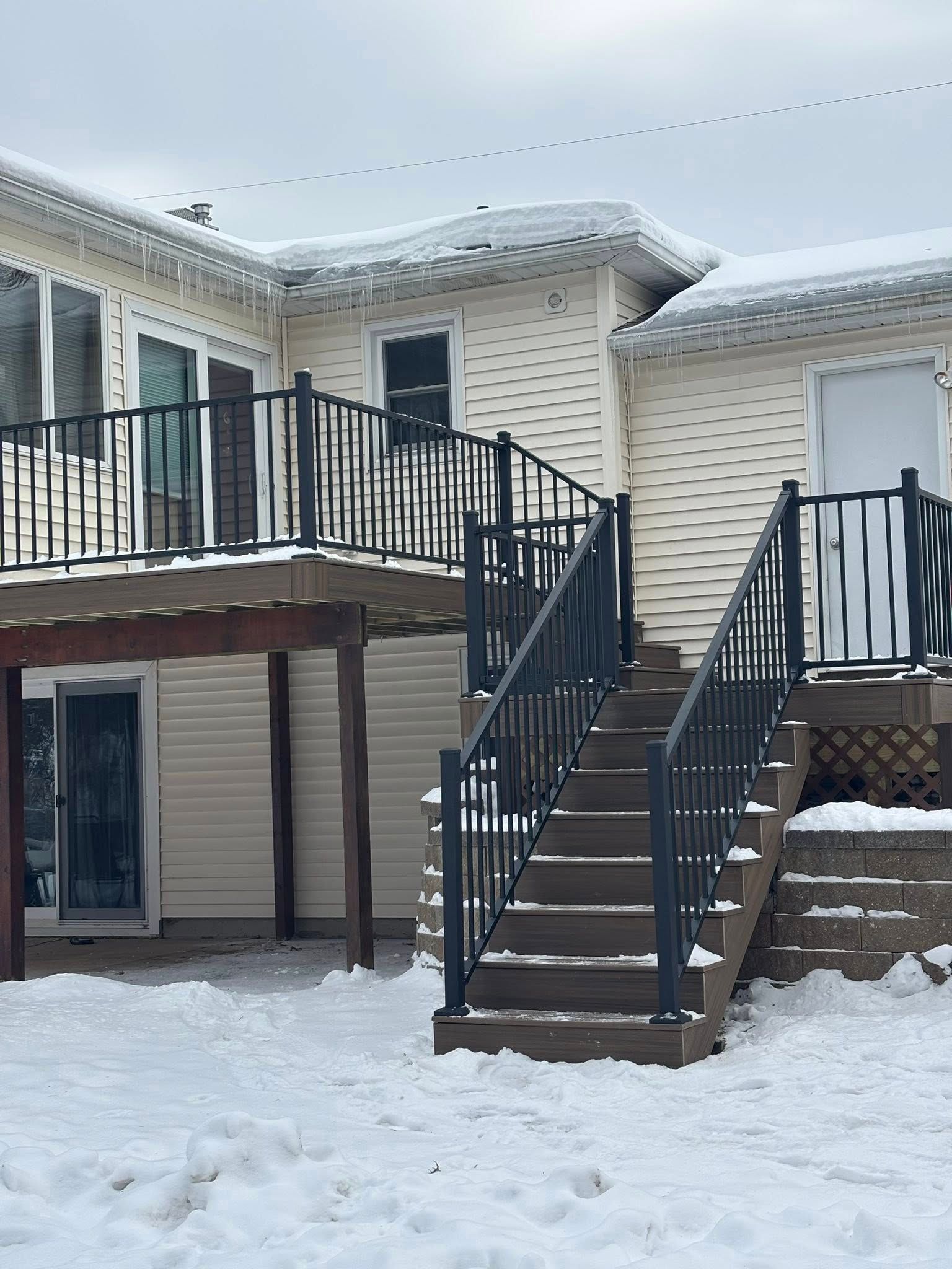 Snowy house exterior with a raised deck, black railings, and stairs leading to the backyard