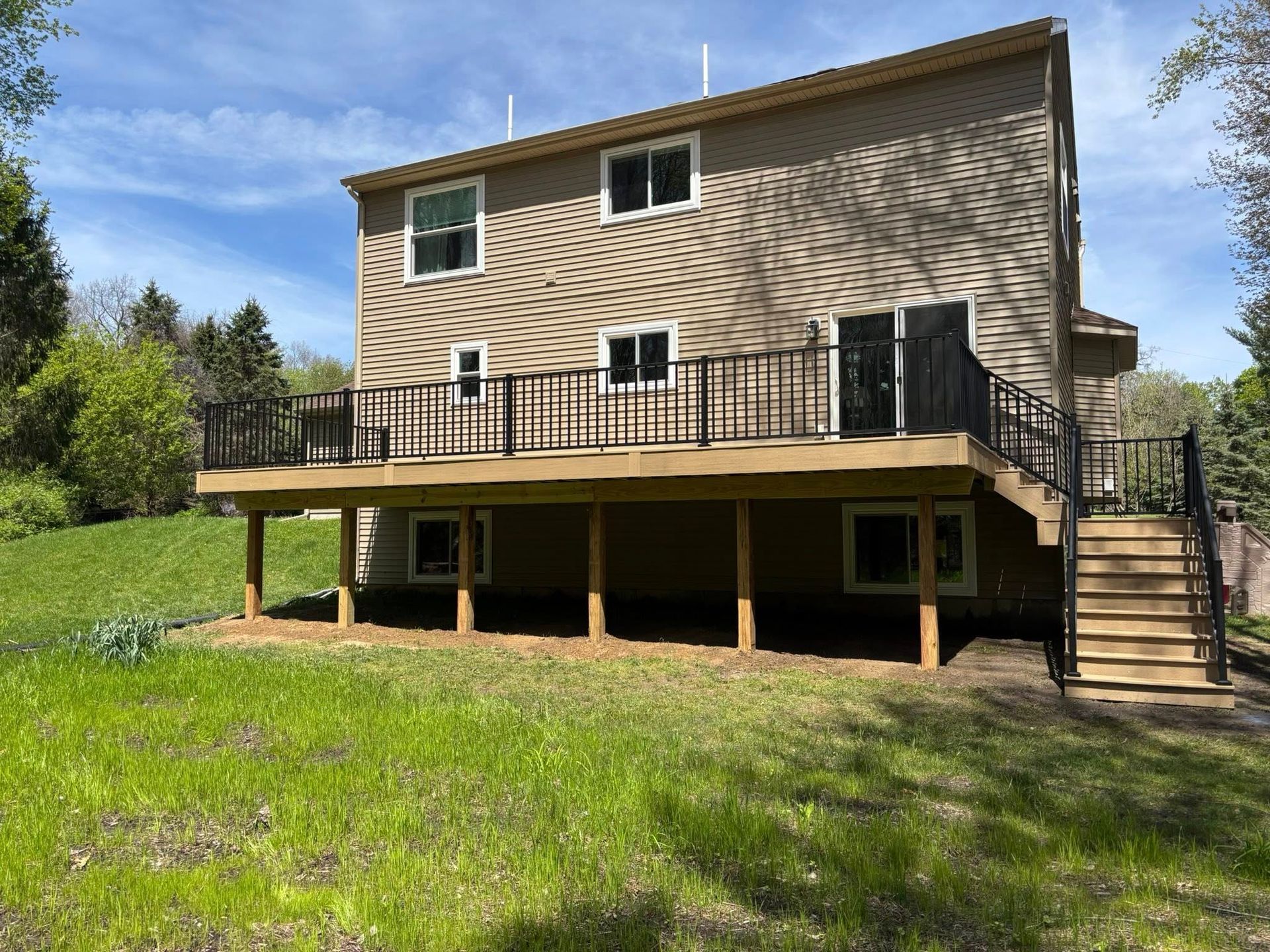 Two-story beige house with a raised deck, stairs, and a grassy backyard on a sunny day