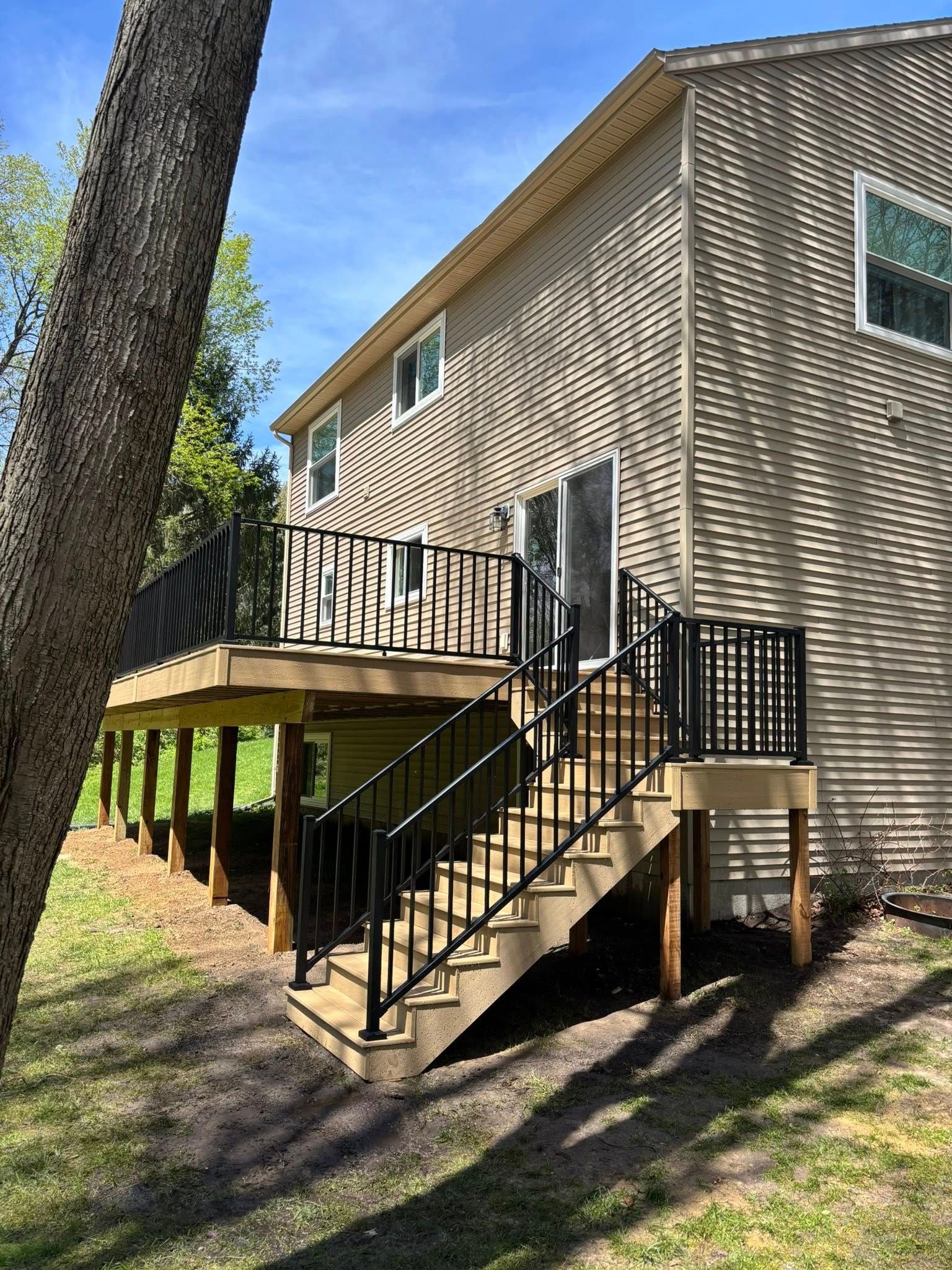 Wooden house exterior with an elevated deck, black stairs, and a tree in the foreground.