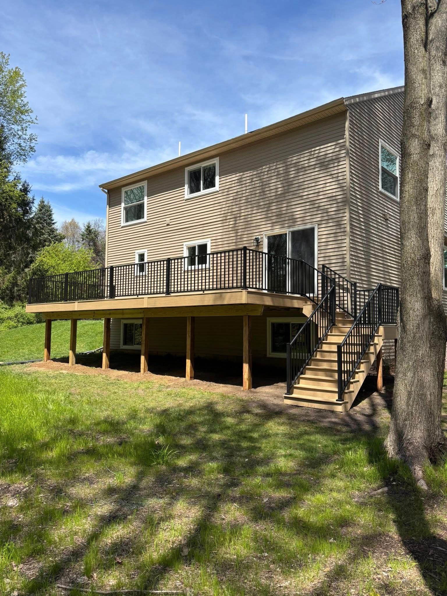 Two-story tan house with a raised wooden deck and stairs in a grassy yard under a blue sky