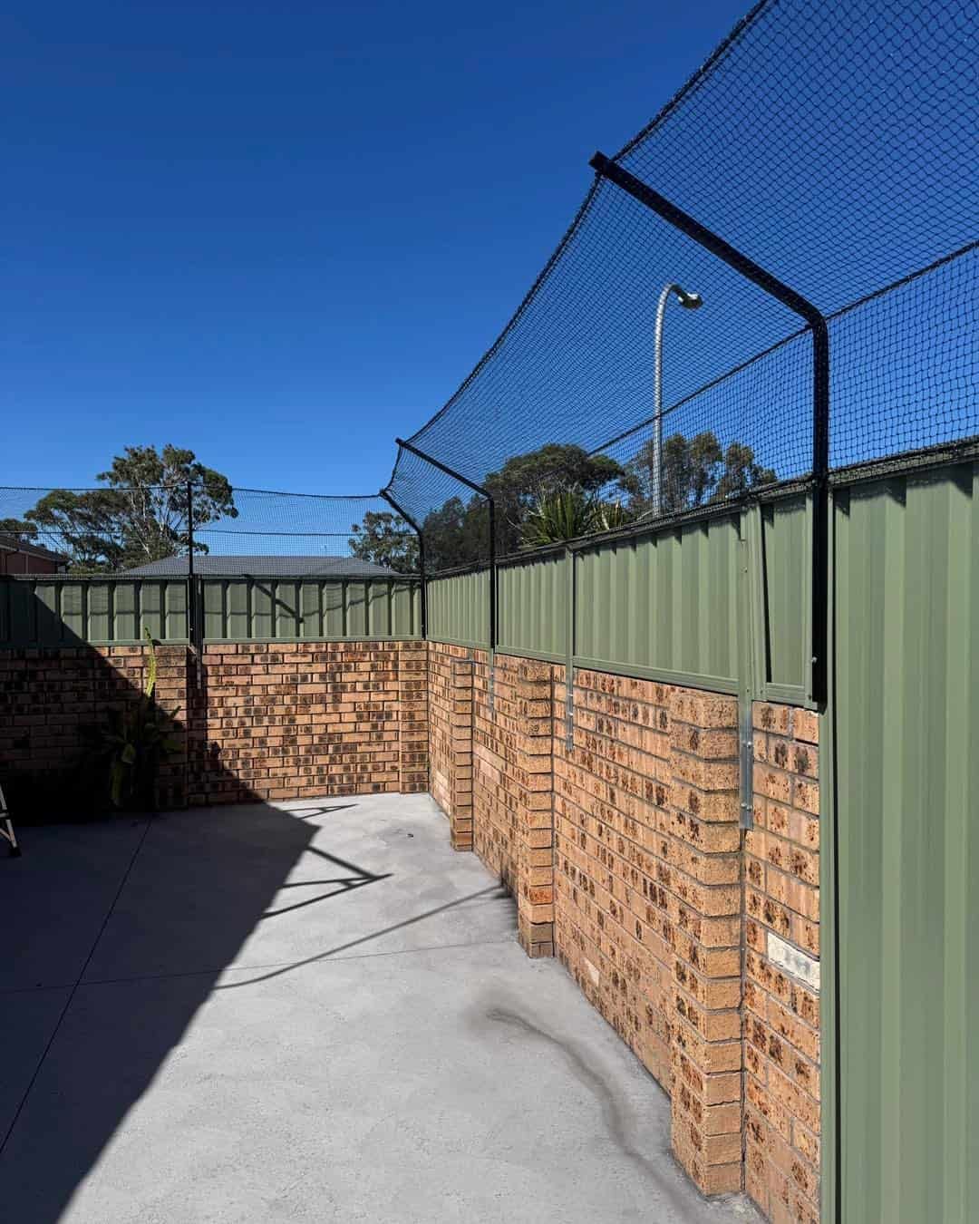 Brick-lined Outdoor Walkway Beside a Green Fence and Tall Netting Under a Clear Blue Sky — Catios By Paulie in Sydney, NSW