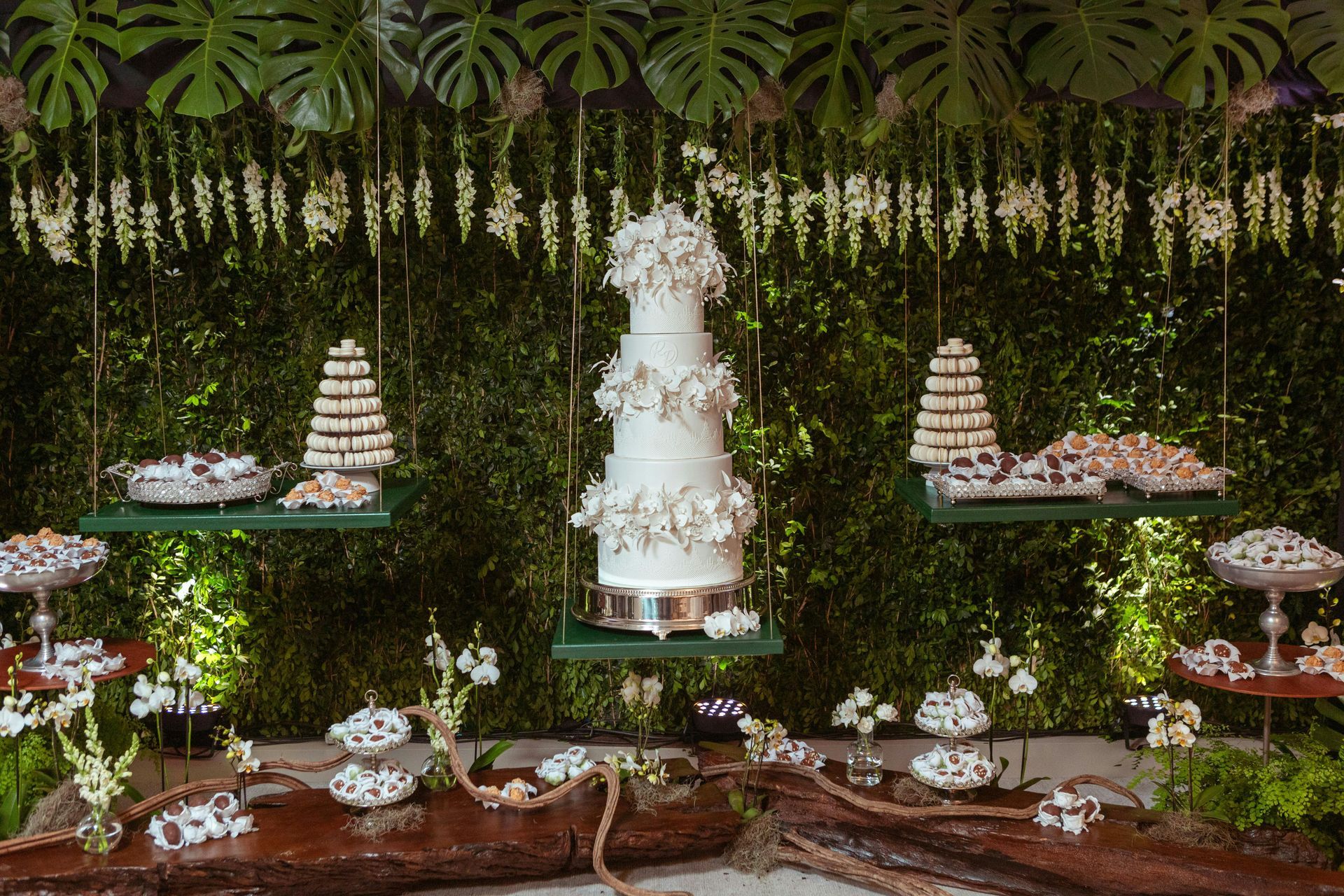 Elegant dessert table with a tall white tiered cake, smaller cakes, and floral decorations under hanging greenery