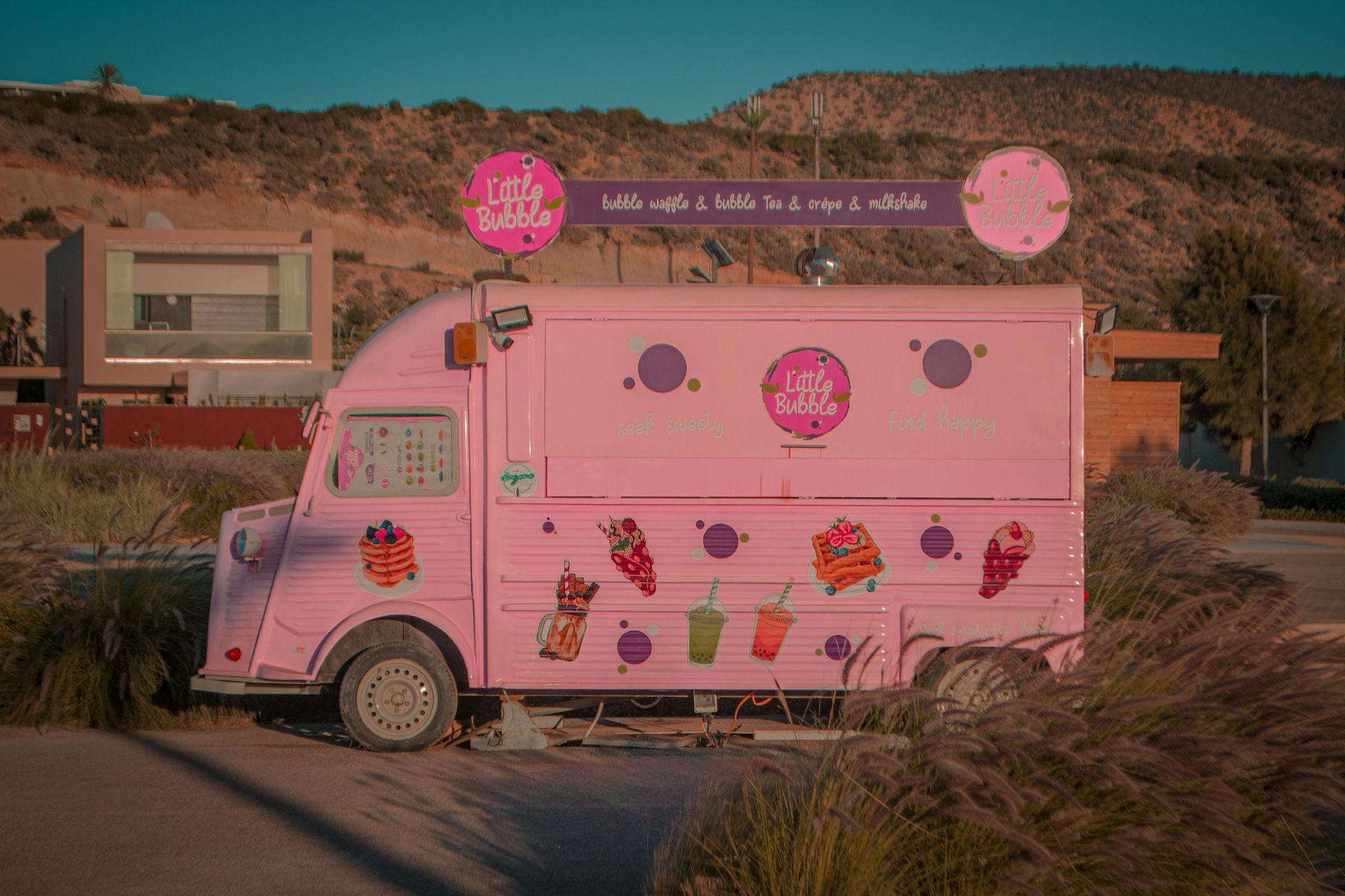 Pink ice cream truck with colorful dessert graphics parked by a roadside in a desert landscape