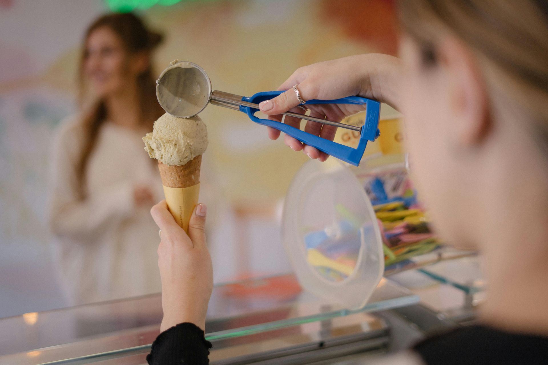 Hand holding an ice cream cone while a blue scoop serves another scoop in an ice cream shop